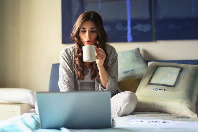 person sitting cross legged on a bed blowing on a cup of coffee while using a laptop